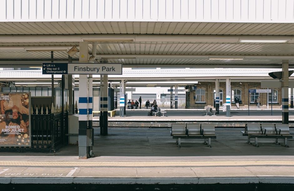 The image shows the Finsbury Park train station platform on a clear day, with a white sign displaying the station name in black letters hanging from the ceiling. Several metal benches are positioned along the platform, some with passengers sitting and waiting. In the background, additional platforms are visible with people walking, some carrying luggage or sitting on benches. The platform is covered by a roof made of metal panels supported by vertical poles, with blue and white safety markings on the poles. A part of the station's brick building can be seen further back, with windows and a timetable board. The platform surface includes tactile paving strips near the edge for safety. The scene depicts a typical urban transport environment suitable for home relocation or furniture transport planning, and occasionally, professional removals services like those provided by Removals Finsbury Park are involved in managing moving logistics and packing activities around such station accesses.