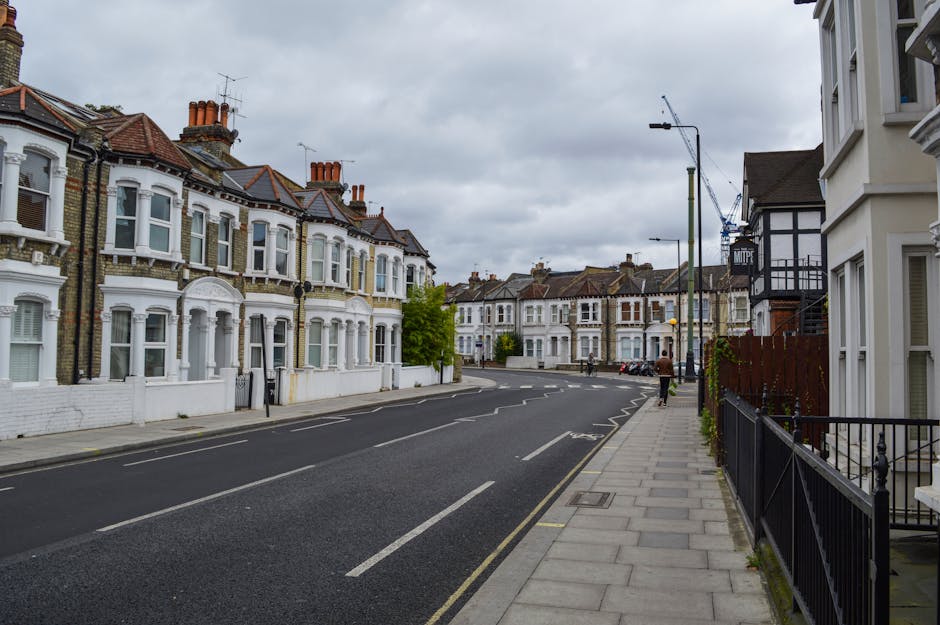 A quiet residential street with terraced Victorian-style houses featuring bay windows, decorative cornices, and brick facades, lined with a paved sidewalk on the right side and a black metal fence in front of one property. The sky is overcast with grey clouds, and there are a few pedestrians walking along the sidewalk. The road has marked lanes and a designated parking area with several parked cars further down. In the background, a construction crane is visible among buildings, indicating ongoing development nearby. The scene captures a typical urban neighbourhood appropriate for home relocation and furniture transport activities, with no visible moving equipment or vehicles present in the image.