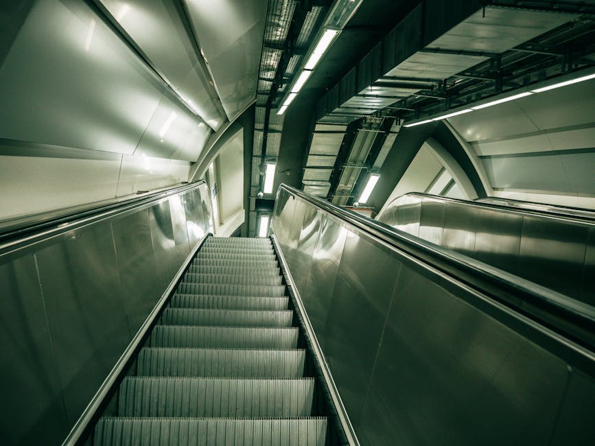A downward-facing view of a modern, metallic escalator located inside a station or building, with stainless steel side panels and steps made of ribbed, anti-slip material. The escalator is situated in a spacious environment with visible structural elements such as beams, support columns, and ceiling fixtures, all illuminated by overhead lighting. To the right, a metallic handrail runs parallel to the escalator, while to the left, a smooth metallic wall extends upward. In the background, there are stairs and what appears to be a platform or transit area, indicating a setting that facilitates home relocation or moving logistics near Finsbury Park Station. The clean, industrial design and lighting suggest a professional environment related to house removals or furniture transport, with the escalator being part of a transport hub used during packing and moving operations.
