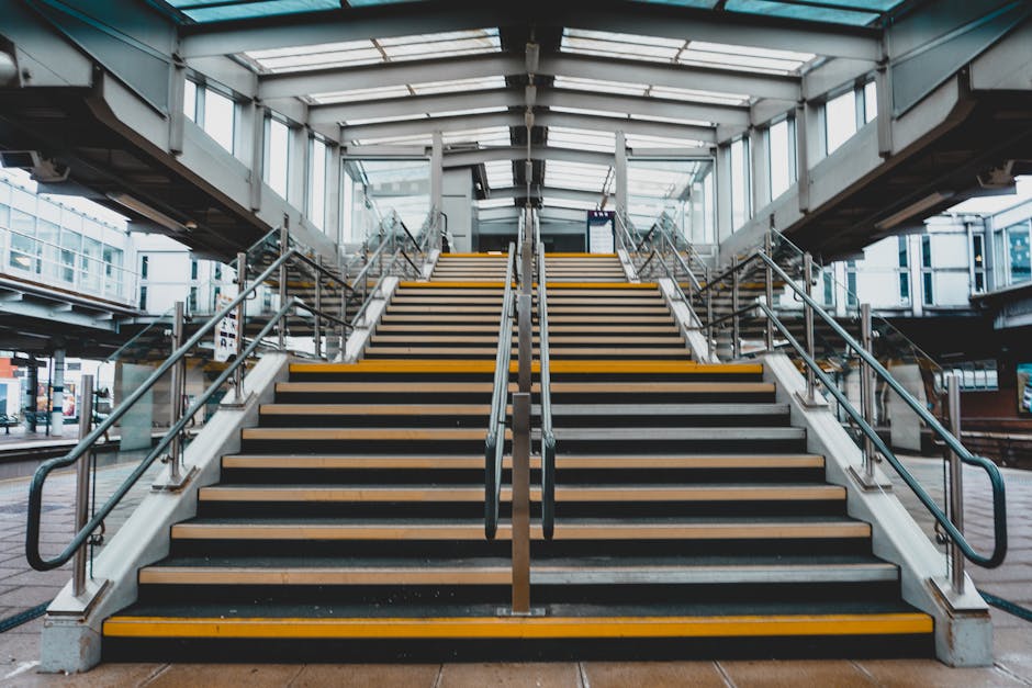 A downward-facing view of a modern, metallic escalator located inside a station or building, with stainless steel side panels and steps made of ribbed, anti-slip material. The escalator is situated in a spacious environment with visible structural elements such as beams, support columns, and ceiling fixtures, all illuminated by overhead lighting. To the right, a metallic handrail runs parallel to the escalator, while to the left, a smooth metallic wall extends upward. In the background, there are stairs and what appears to be a platform or transit area, indicating a setting that facilitates home relocation or moving logistics near Finsbury Park Station. The clean, industrial design and lighting suggest a professional environment related to house removals or furniture transport, with the escalator being part of a transport hub used during packing and moving operations.