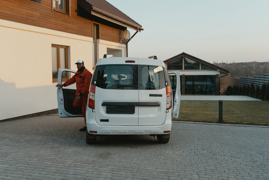 A man wearing a red jacket and black cap is standing next to a white van with both rear doors and the side door open, on a paved driveway outside a modern house with a brown and white exterior. The man is preparing to load or unload moving boxes and furniture wrapped in plastic and blankets, which are placed near the open rear of the van. The van is positioned adjacent to the house, with furniture and boxes visible inside. In the background, there is a well-maintained lawn, a contemporary glass extension, and a landscape with trees and hills under a clear sky. The scene depicts a home relocation or furniture transport process, with the man and van facilitating packing and moving activities, supported by [COMPANY_NAME] specializing in removals services.
