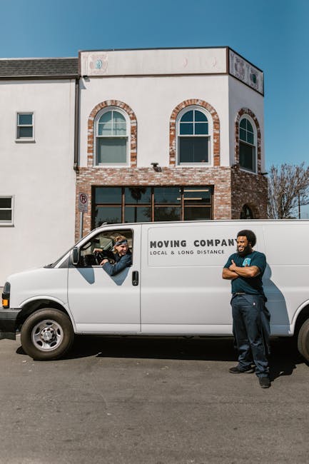 A white moving van parked on a street in front of a modern, two-storey building with a brick and white exterior, featuring arched windows on the upper floor. The van, labelled with the words 'MOVING COMPANY LOCAL & LONG DISTANCE', is being interacted with by two men wearing casual clothing; one inside the driver’s seat, speaking through the window, and the other standing outside the van with arms crossed, smiling. The scene is set during daytime with clear blue skies, and the men appear engaged in a conversation related to the home relocation process, which may involve packing, loading, or transporting furniture and moving boxes as part of a professional removals service provided by [COMPANY_NAME]. The environment suggests preparations for a house move, with the van positioned close to the building entrance, possibly indicating active loading or unloading for a house removal or furniture transport joblinked to the services on the webpage [PAGE_TITLE].