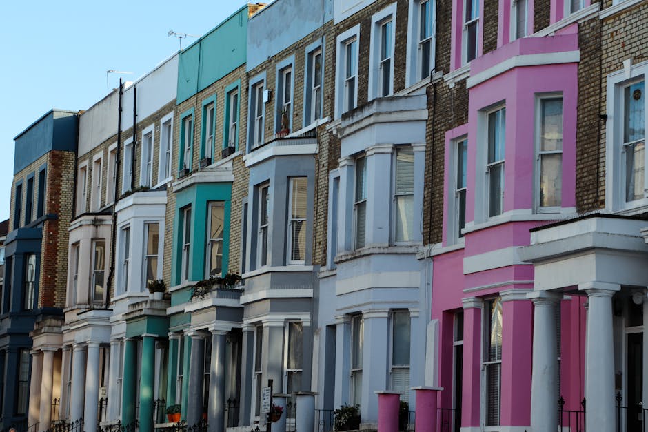 A row of Victorian terraced houses in Stroud Green, Finsbury Park, featuring pastel-colored facades including shades of blue, turquoise, white, cream, and pink. The buildings have bay windows with white frames and small front gardens with decorative iron fencing. The street is shown during daylight with clear weather, showcasing the uniform architectural style with slight variations in color and window placement. The image depicts a typical residential area in Finsbury Park, suitable for home relocation or furniture transport services, with the houses closely positioned along the pavement, emphasizing the urban setting for moving and packing processes. Removals Finsbury Park specializes in house removals, and this visual context supports their professional moving and packing services for Victorian terrace properties.